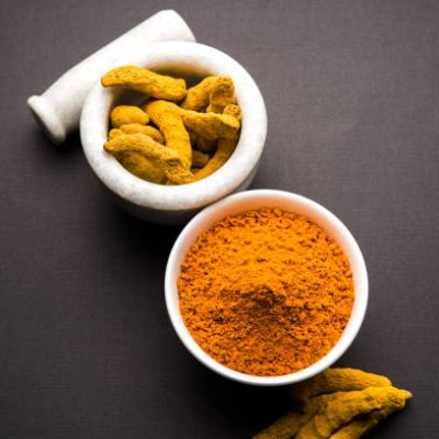 Turmeric powder in ceramic bowl with raw dried turmeric over plain background
